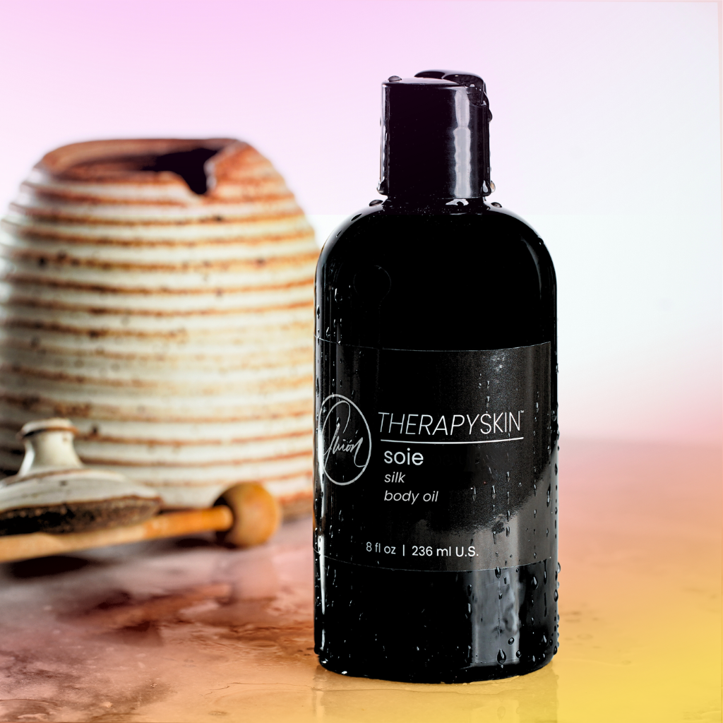 A bottle of THERAPYSKIN Silk Body Oil, with a honey jar and dipper in the background.